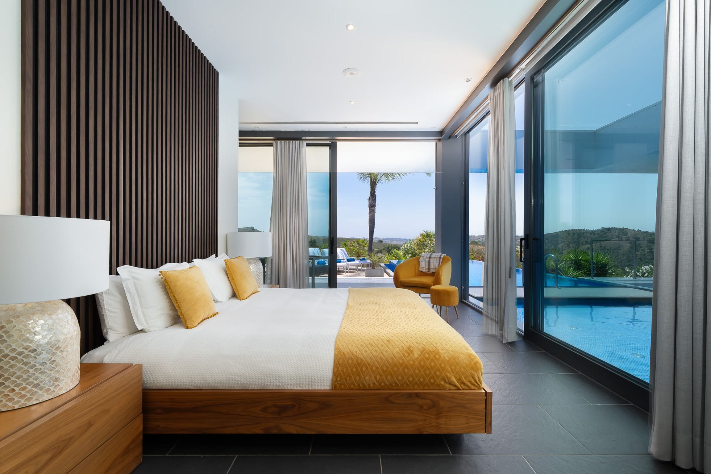 Bedroom with a floating walnut bed, ochre throw and full-height glass opening to the pool and hills