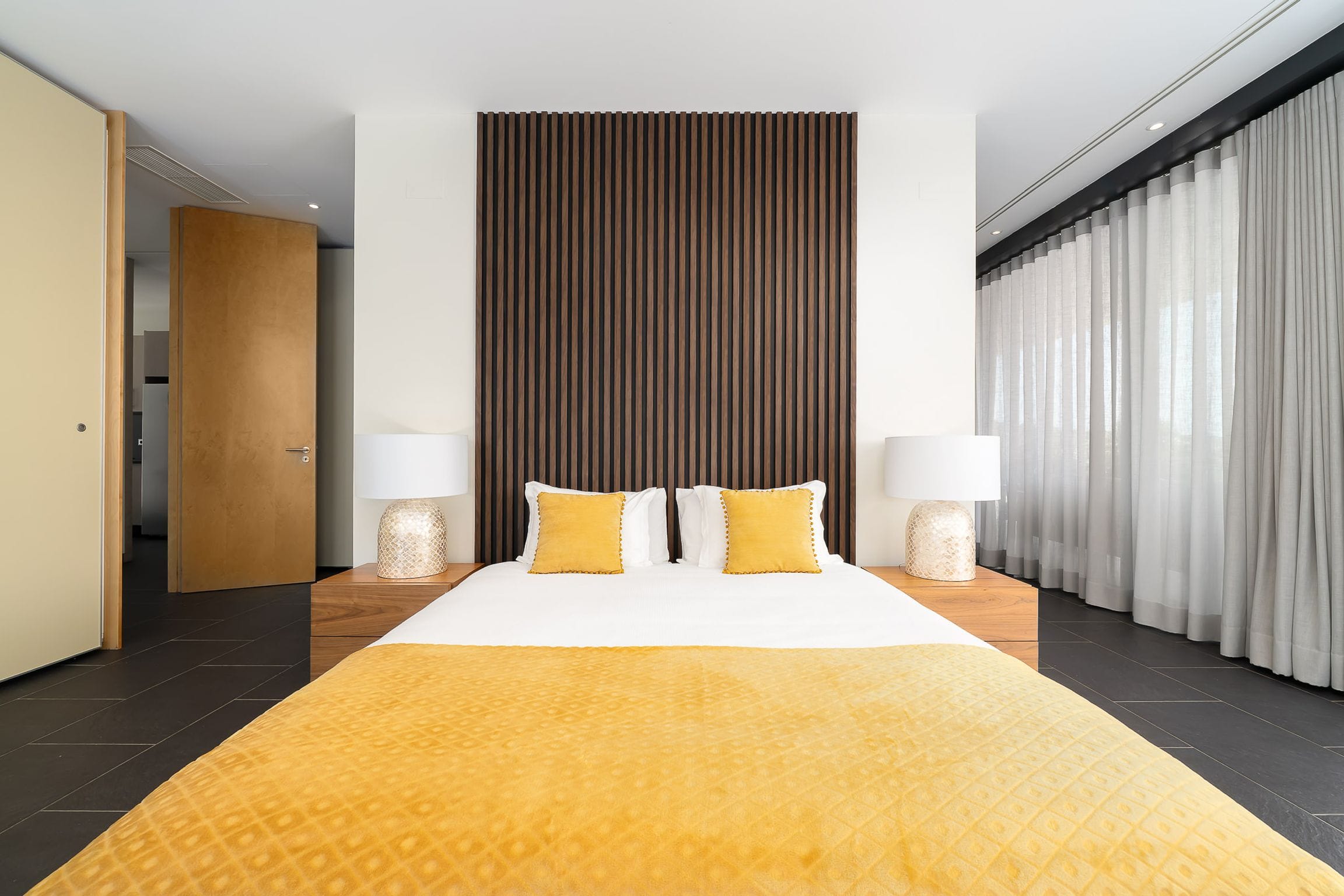 Symmetrical bedroom with a slatted timber headboard wall, ochre bedding and sheer curtains